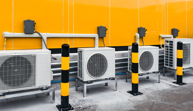 Air conditioning units installed against a bright yellow wall, featuring multiple outdoor compressors with protective bollards, illustrating L&S Air LLC's HVAC installation services in Hidalgo County.
