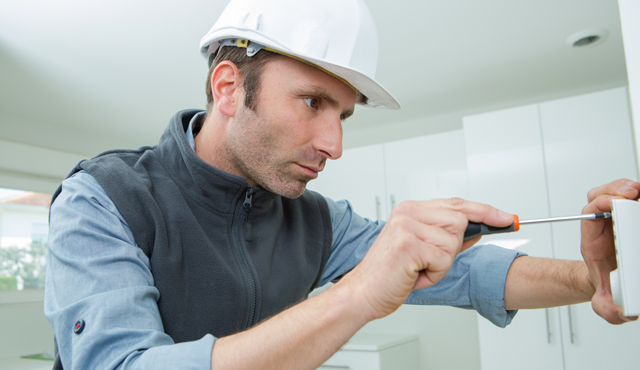 Technician installing a thermostat with a screwdriver, wearing a hard hat, in a modern indoor setting, emphasizing professional installation services by L&S Air LLC for improved HVAC efficiency.